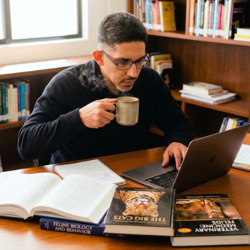 Faustino Ribeiro concentrado a estudar num portátil, com livros sobre biologia felina na secretária e uma chávena de café, ambiente de pesquisa focado.